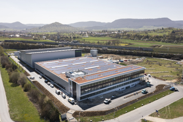 Luftaufnahme eines großen Gebäudes mit Solarmodulen auf dem Dach, umgeben von Fahrzeugen, Bäumen, Gras, Pfählen und einer klaren blauen Himmel; das Gebäude ist die erste solarbetriebene Fabrik Deutschlands.