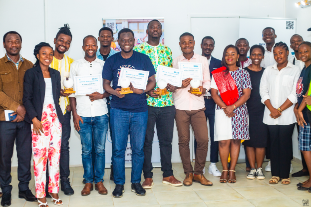 Eine Gruppe von Menschen, die zusammen in einem Raum stehen, lächeln und Zertifikate halten, mit einem Tisch mit Gegenständen rechts und Tafeln und einem Plakat im Hintergrund, feiern die 2019 WIK Awards.