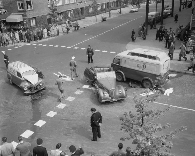 Schwarz-weißes Bild eines Autounfalls an einer Straßenkreuzung mit mehreren Fahrzeugen, darunter ein Lieferwagen, und einer Gruppe von Schaulustigen; Gebäude, Bäume, Pfosten und Texttafeln sind im Hintergrund zu sehen.