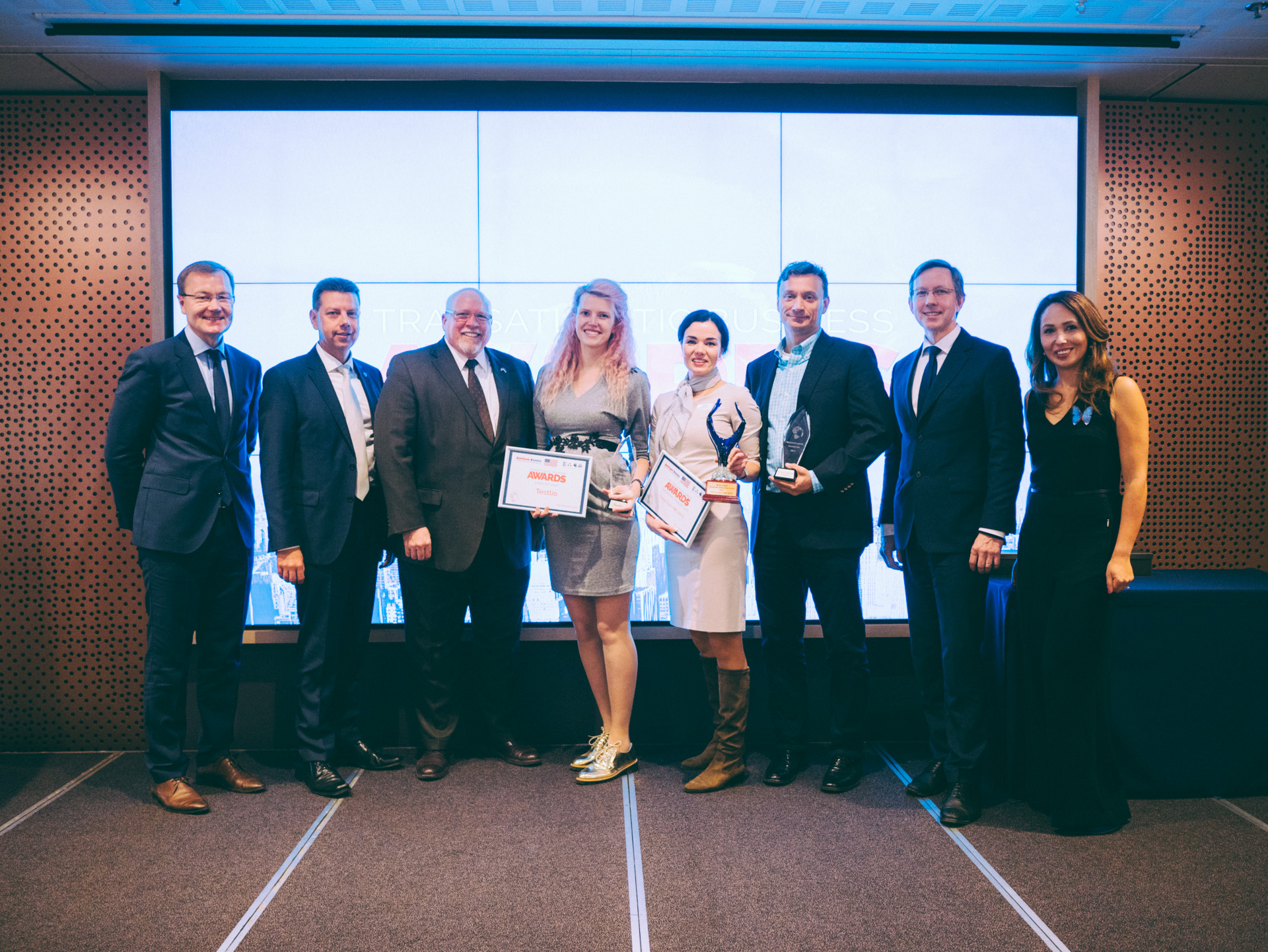 Eine Gruppe strahlender Menschen hält Zertifikate in der Hand und steht zusammen vor einem Bildschirm, mit einem rechts daneben stehenden Tisch, der mit einer schwarzen Decke bedeckt ist und einem Gegenstand an der Wand im Hintergrund, feiern ihren Sieg bei den International Business Awards 2018.