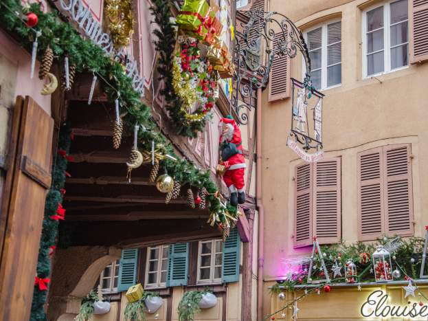 Ein Gebäude in Colmar, Frankreich, geschmückt mit Weihnachtsdekorationen wie Girlanden, Lichtern und einer Santa-Claus-Figur, mit einem Schild, auf dem "Colmar Weihnachtsmarkt" steht.