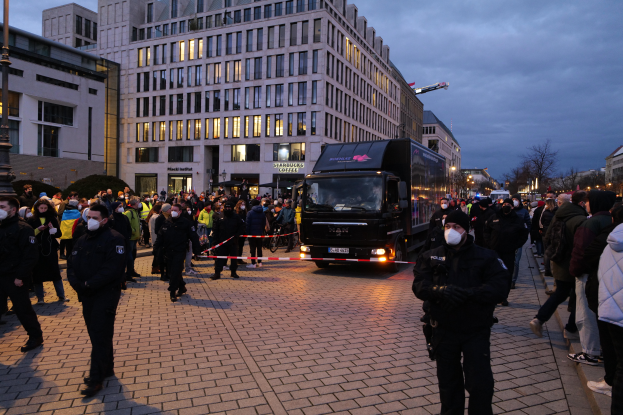 Eine Gruppe von Menschen steht vor einem Lastwagen auf einer Straße, umgeben von Gebäuden, Laternen, Bäumen und einem bewölkten Himmel, wobei einige Mützen und Masken tragen und ein Band mit einem Pfahl im Vordergrund.