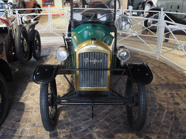 Ein Oldtimer in einem Museum ausgestellt, umgeben von anderen Autos und einem Geländer, mit Informationsschildern im Hintergrund.
