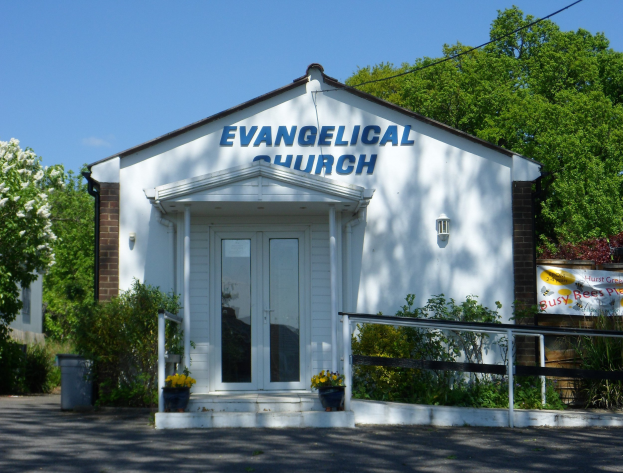 Evangelische Kirche mit Straße im Vordergrund, umgeben von Grünfläche unter einem klaren blauen Himmel.