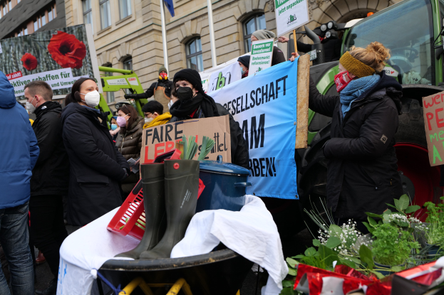 Eine Gruppe von maskierten Menschen mit Protestschildern vor einem Lastwagen, mit einem Tisch, Pflanzen und Gebäuden im Hintergrund.
