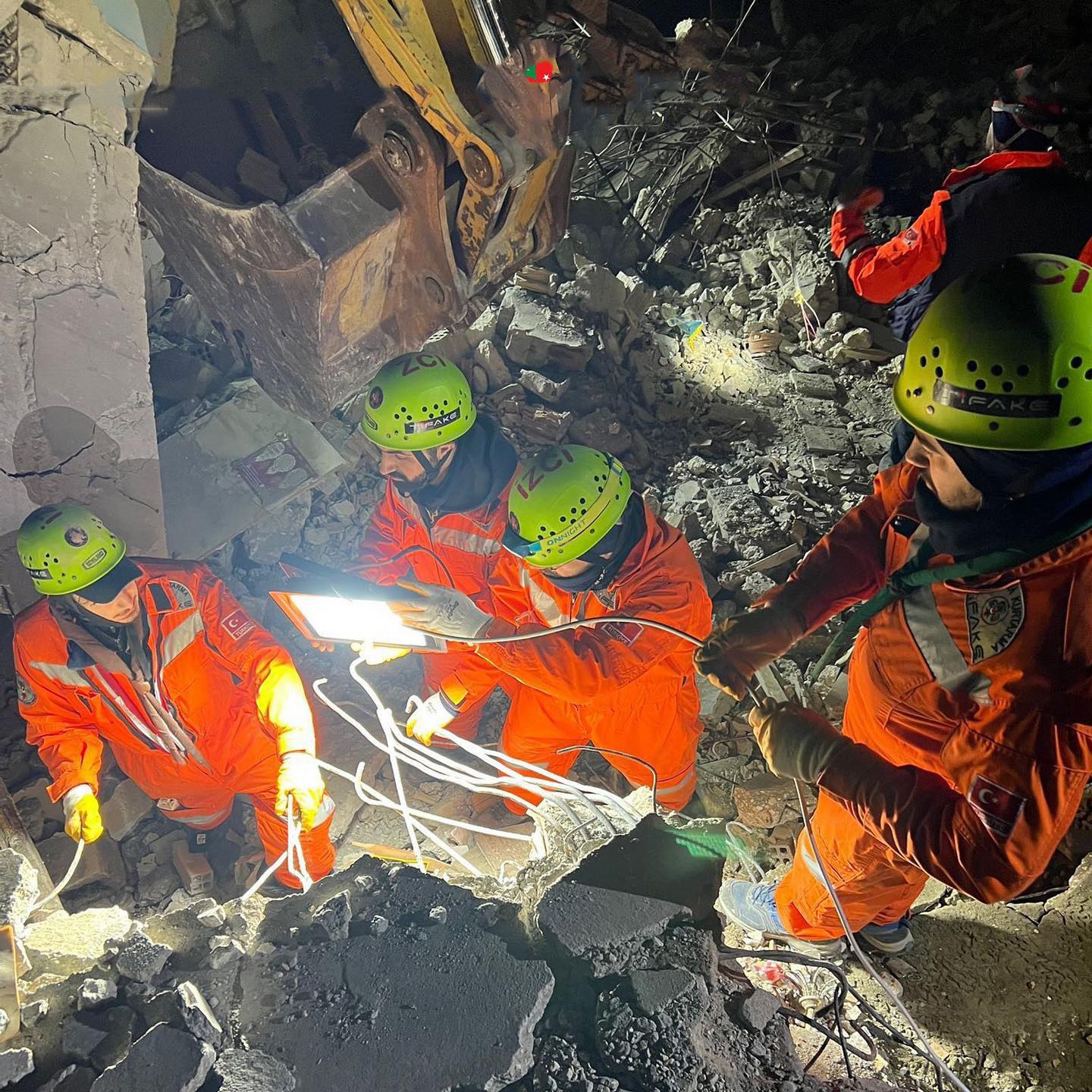 Gruppe von Rettern in orangefarbenen Uniformen und grünen Helmen beseitigen Trömer von einem eingestürzten Gebäude mit einem Kran im Hintergrund.