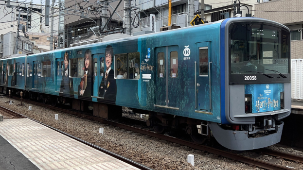 Blauer Zug fährt auf Schienen neben einem Bahnsteig mit Strommasten und Drähten, Gebäude und klarer Himmel im Hintergrund.