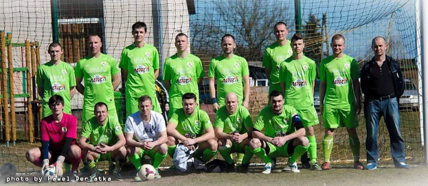 Gruppe von Männern in grünen T-Shirts auf einem Fußballfeld mit einem Ball davor, einige in der Hocke, in der Nähe eines Torpfostens mit Bäumen, Gebäuden und Fahrzeugen im Hintergrund.