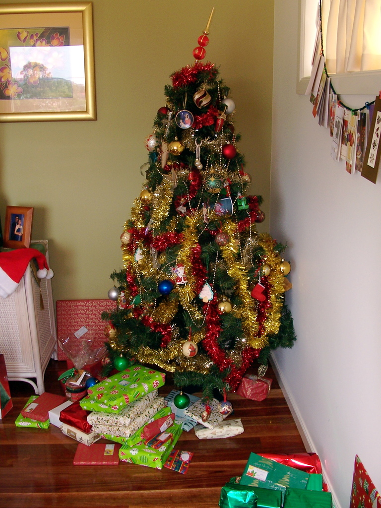 Ein geschmückter Weihnachtsbaum mit Geschenken darunter, ein Foto auf der linken Wand und ein Vorhang am rechten Fenster.