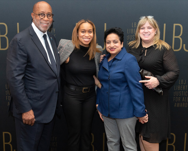 Vier Personen stehen zusammen auf einem roten Teppich, lächeln und posieren für ein Foto, mit einem 'Liberty Justice for All Awards'-Schild im Hintergrund.