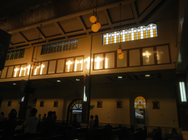 Das Innere einer Kirche mit Menschen auf Bänken und einigen Stehenden, Wände mit Fenstern geschmückt und Lampen von der Decke hängend.