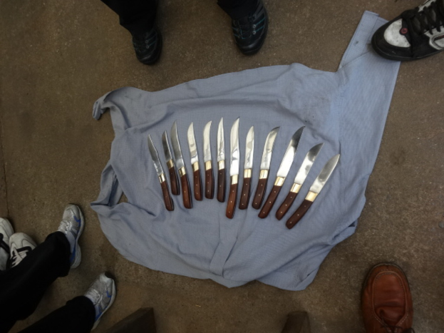 Eine Sammlung von Messern, die auf einem Tuch auf einer Straße ausgebreitet sind, mit zahlreichen Menschenbeinen drumherum.