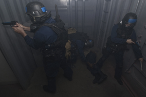 Eine Gruppe von Polizisten in Einsatzausrüstung, einer mit einer Waffe, stehend neben einem Metallbehälter und einer Leiter in einem begrenzten Raum.