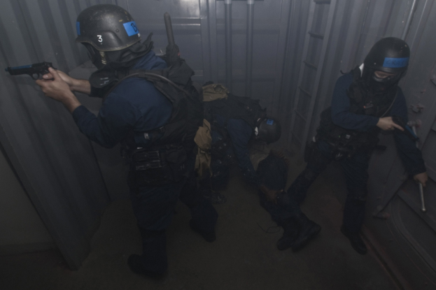 Eine Gruppe von Polizisten in Einsatzausrüstung, einer mit einer Waffe, stehend neben einem Metallbehälter und einer Leiter in einem begrenzten Raum.