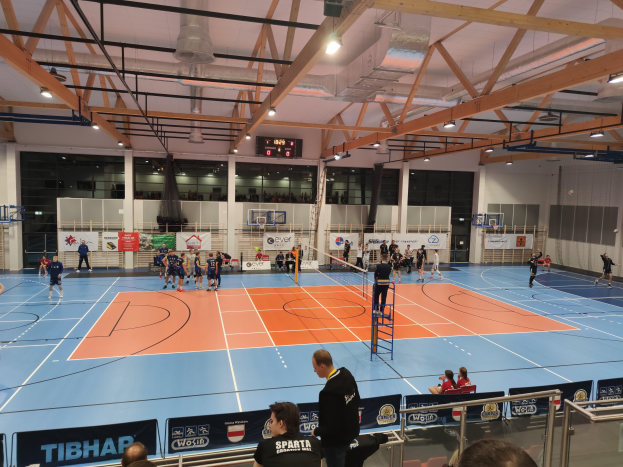 Gruppe von Menschen, die Handball in einer Turnhalle spielen, mit Zuschauern, einer Leiter, Geländern, Tafeln mit Text, Wänden, Säulen, einem Bildschirm und Deckenleuchten.
