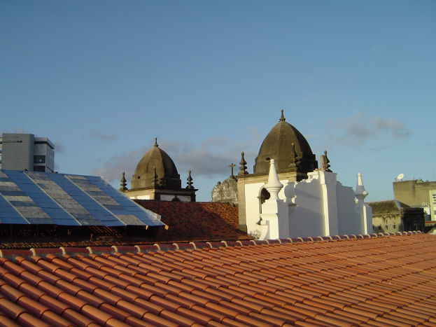Stadtansicht mit mehreren Gebäuden im Vordergrund und einem klaren blauen Himmel im Hintergrund, die Solarpanels auf dem Dach eines Gebäudes zeigen.