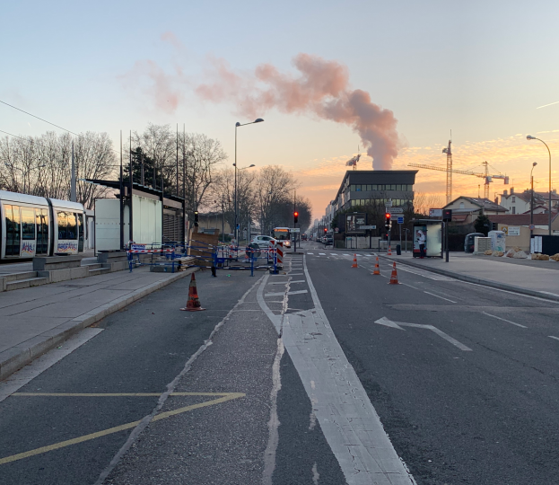 Eine Stadtstraße mit Verkehrskegeln, einem Bus, Laternenmasten, Bäumen, Gebäuden, Kränen und Rauch, der aus einem entfernten Gebäude aufsteigt.