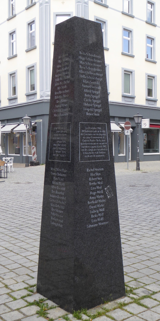 Ein Denkmal mit Inschrift steht in einem Stadtplatz umgeben von Gebäuden, Straßenmöbeln und Menschen unter einem sichtbaren Himmel.