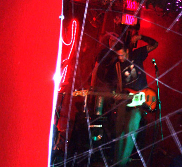 Ein Mann in einem schwarzen T-Shirt steht in der Mitte und hält eine Gitarre, während ein anderer Mann hinter ihm möglicherweise Instrumente spielt, unter hellen Scheinwerfern und vor einer roten Wand bei einem Musikkonzert.