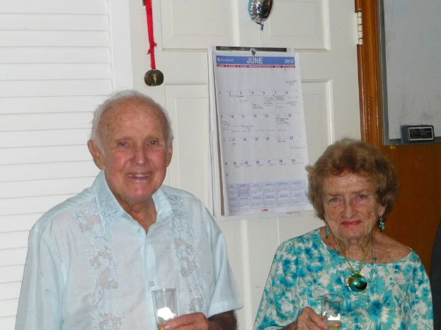 Ein älterer Mann in einem Hemd hält ein Glas und lächelt, neben einer Frau in einem blauen Kleid mit Ohrringen und einer Kette, die ebenfalls ein Glas hält, mit Medaillen und einem Kalender im Hintergrund.