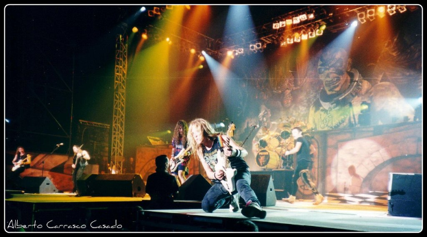 Eine Gruppe von Menschen, die auf einer Bühne Musikinstrumente spielen, mit Lichtern oben und auf der Bühne, Graffiti an der Wand, einer Person in der Nähe der Bühne und Text auf dem Bild.