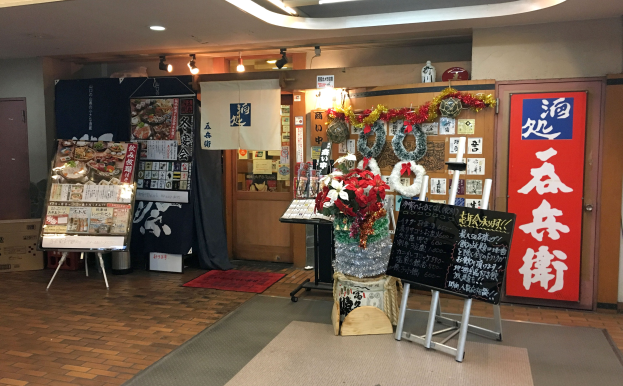 Ein unordentlicher Raum mit verschiedenen Gegenständen, darunter eine Blumenvase, ein Textbrett auf einem Ständer, ein Teppich, eine Kartonbox, Papiere an der Wand, Regalen mit Gegenständen, ein Vorhang, eine Tür, Deckenleuchten und ein Schild, das den Eingang eines japanischen Restaurants vermuten lässt.