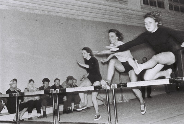 Zwei Frauen in der Luft beim Überwinden eines Hindernisses in einer Turnhalle mit Zuschauern und Fenstern im Hintergrund.