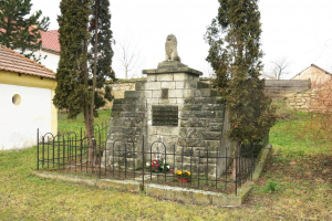 Ein steinernes Denkmal auf einer grünen Wiese, umgeben von einem Metallzaun und geschmückt mit Blumensträußen, mit Bäumen, Häusern und einer Wand im Hintergrund unter einem klaren blauen Himmel dient als Holocaust-Gedenkstätte in Sibiu, Rumänien.