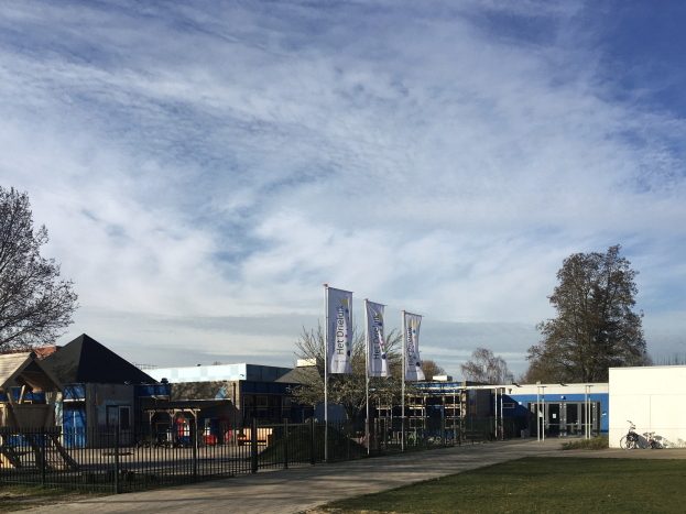 Großes Gebäude mit einem grasbewachsenen Bereich davor, umgeben von einem Metallzaun, mit einem Weg, Bäumen, Pfählen, Bannern, einem geparkten Fahrrad, nahen Häusern und einem bewölkten Himmel, identifiziert als der neue Spielplatz der Schule.