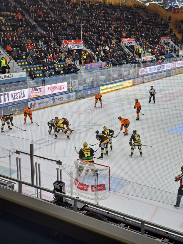 Eine Gruppe von Menschen, die Hockey auf einem Eisstadion spielt, mit einem Netz, einer Umzäunung, Zuschauern auf Stühlen und beleuchteten Bannern im Hintergrund.