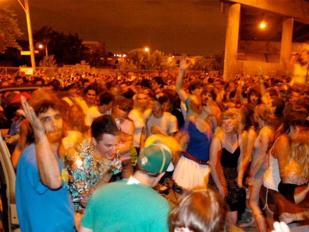 Menschen tanzen im Vordergrund eines Konzerts, mit Lichtern, Pfählen, Bäumen und anderen Objekten im Hintergrund.