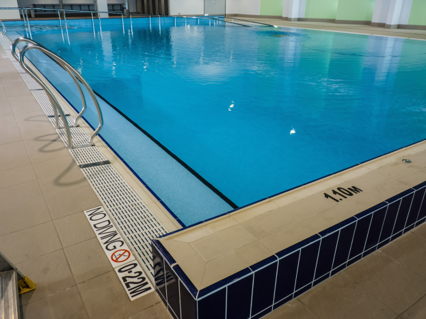 Großes Hallenbad in einem geräumigen Gebäude mit gefliestem Boden, Metallträgern, einer Schautafel, einer Tür, einer Wand und Deckenbeleuchtung.