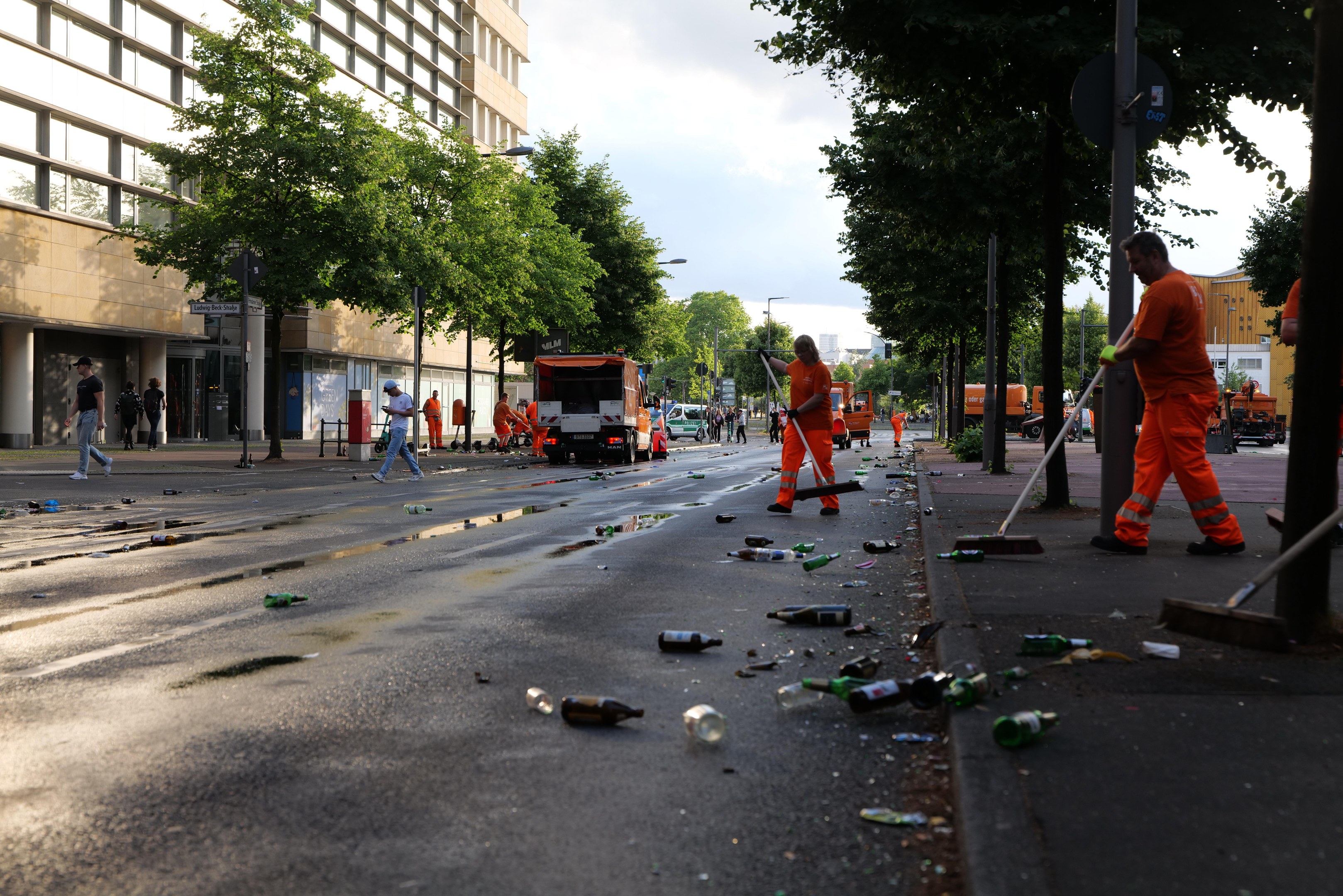 Eine Gruppe von Menschen in orangefarbigen Uniformen, die Müll von einer Straße mit verstreuter Abfall, Bäumen und Fahrzeugen, sowie Gebäuden und bewölktem Himmel im Hintergrund sammeln.