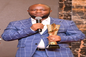 Ein Mann in einem blauen Anzug hält ein Mikrofon und eine Trophäe und steht selbstbewusst mit einem fröhlichen Gesichtsausdruck vor einer Wand.