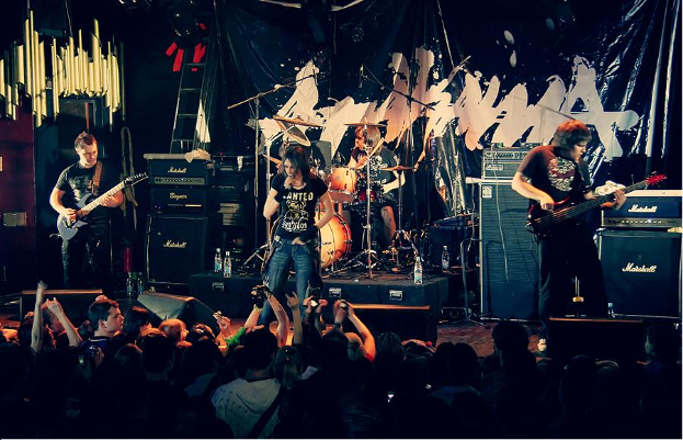 Eine Gruppe von Musikern, die auf der Bühne mit Gitarren und Trommeln spielen, Mikrofone und Ständer sowie ein Banner im Hintergrund und Text am unteren Rand mit der Aufschrift "The Wanted - Live at the O2 Academy, London".
