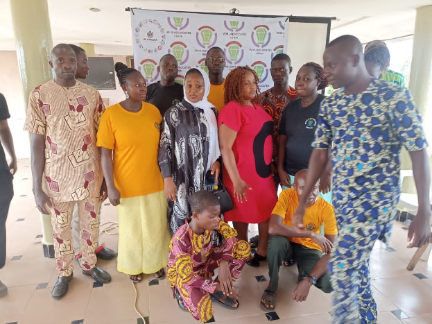 Eine Gruppe von Menschen steht vor einer Fahne mit der Aufschrift "Advocates in Ghana: Ein Schritt-für-Schritt-Führer", mit Stühlen, Säulen, einem Fernseher und Deckenleuchten im Hintergrund.