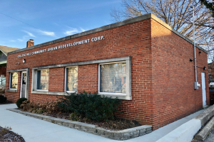 Ein Ziegelgebäude mit einer Tafel, die "College Hill Community Urban Redevelopment Corp." trägt, umgeben von Pflanzen, einem Weg, Bäumen, Häusern, einem Straßenmast, Drähten und einem bewölkten Himmel.