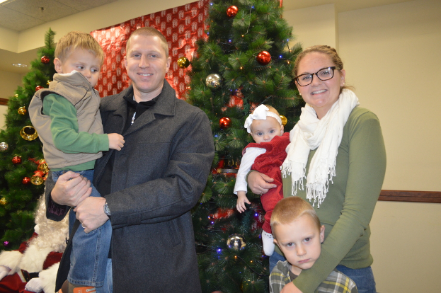 Ein Mann, eine Frau und Kinder stehen im Vordergrund mit Weihnachtsbumen, Santa und einem Plakat im Hintergrund.