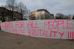 Eine Gruppe von Menschen, die auf dem Boden stehen und eine Fahne halten, auf der steht "Rechte für alle Menschen Stoppt Polizeigewalt", mit einem Straßenschild, einem Hinweisschild, Bäumen, Gebäuden mit Fenstern und einem bewölkten Himmel im Hintergrund.