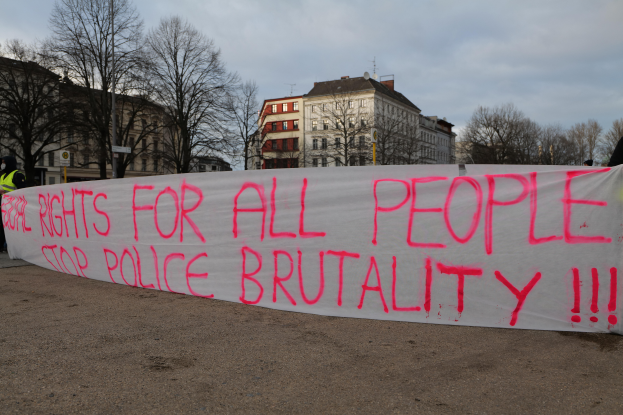 Eine Gruppe von Menschen, die auf dem Boden stehen und eine Fahne halten, auf der steht "Rechte für alle Menschen Stoppt Polizeigewalt", mit einem Straßenschild, einem Hinweisschild, Bäumen, Gebäuden mit Fenstern und einem bewölkten Himmel im Hintergrund.