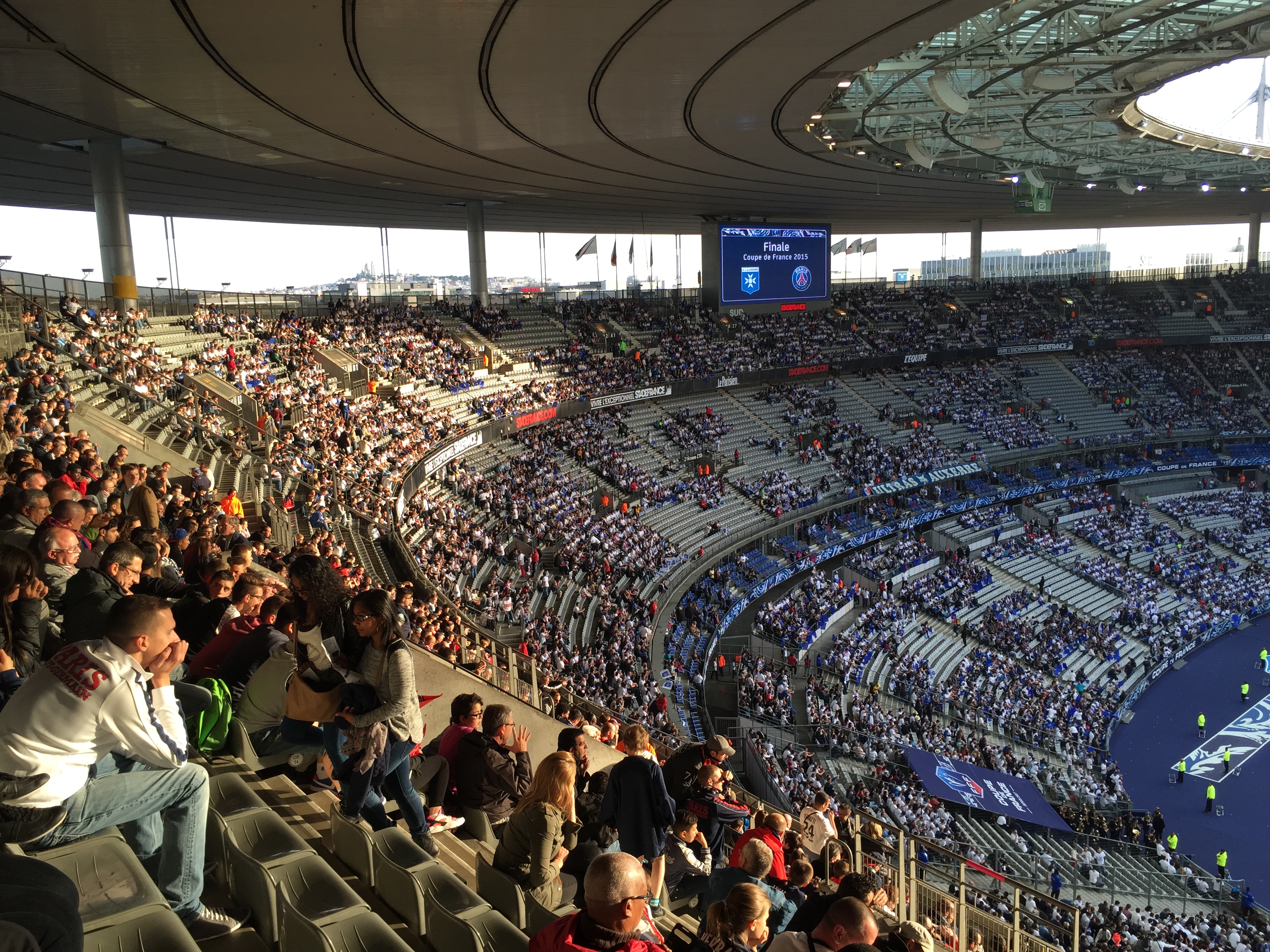 Eine Menschenmenge sitzt in einem Stadion und schaut ein Fußballspiel, mit einer Bühne auf der rechten Seite, Fahnen, Stangen und einem Bildschirm im Hintergrund, unter einem sichtbaren Himmel, im Allianz Stadion in München, Deutschland.