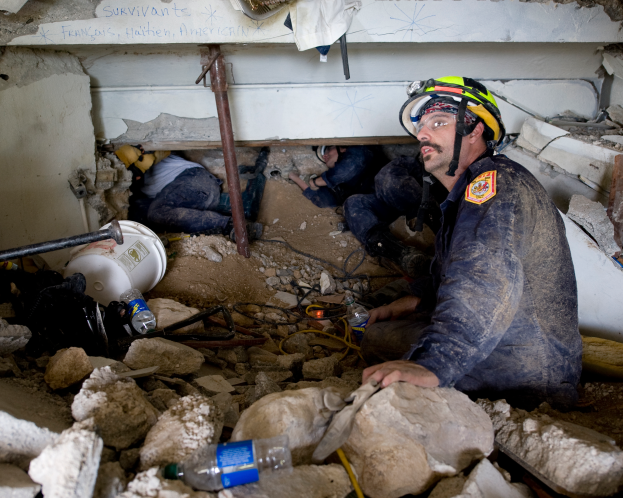 Ein Feuerwehrmann in einem Helm sitzt zwischen Trümmern, darunter Steine und Flaschen, mit einer Wand im Hintergrund, auf der ein Text steht.