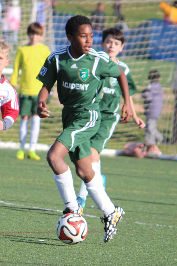 Zwei Personen spielen Fußball in einem Stadion mit wenigen Zuschauern und einem Netz im Hintergrund.