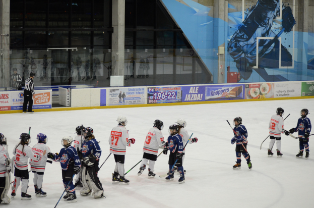 Gruppe junger Menschen beim Eishockey auf einer Indoor-Eisfläche, mit Helmen und Stöcken, einer Person auf der linken Seite und Bannern, Säulen und Glaswänden im Hintergrund.