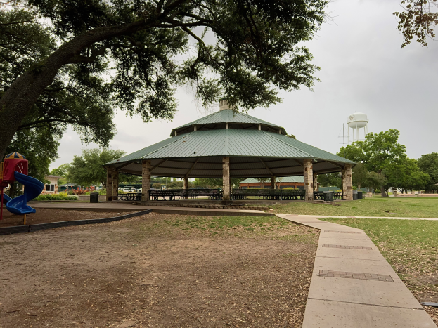 Ein Park mit einem Pavillon in der Mitte, umgeben von Bäumen, Bänken und einer Rutsche, mit einem Weg, der zum Pavillon führt, und einem Wassertank im Hintergrund unter einem klaren blauen Himmel.