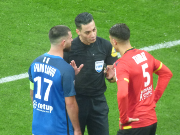 Schiedsrichter spricht mit zwei Männern auf einem Fußballfeld; der Mann in der Mitte trägt ein schwarzes Hemd und Hosen, der Mann links trägt ein blau-rotes T-Shirt mit Text und Brille, beide Männer tragen Brillen.