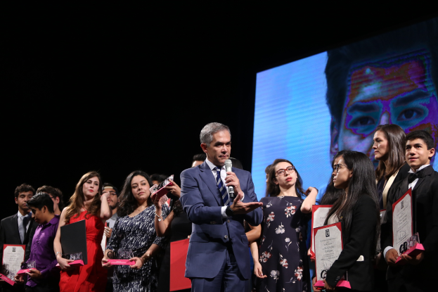 Ein Mann im Anzug und mit Krawatte hält ein Mikrofon auf der Bühne, umgeben von Menschen mit Auszeichnungen, mit einem Bildschirm im Hintergrund.