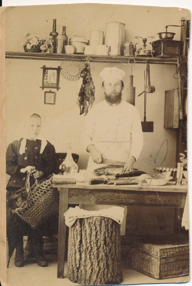 Ein altes Foto eines Mannes in einer Küche mit einem Messer in der Hand und einer Frau auf einem Stuhl mit einem Korb, umgeben von verschiedenen Gegenständen auf dem Tisch und gerahmten Fotos an der Wand.