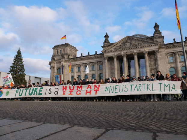 Gruppe von Menschen mit einem Transparent 'Zukunft ist ein Menschenrecht' vor dem Reichstag in Berlin, mit Bäumen und Fahnenmasten im Hintergrund.