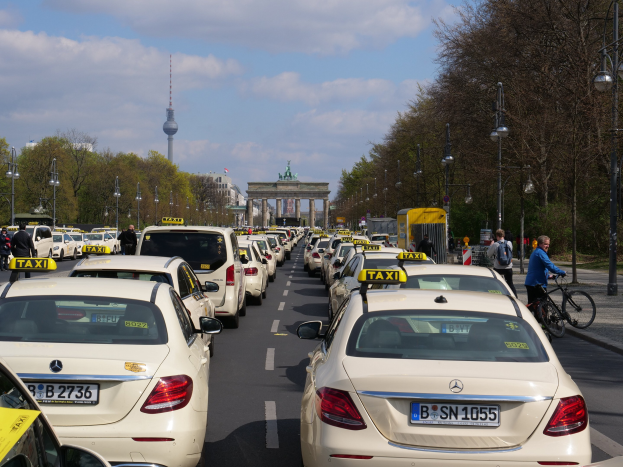 Eine lange Reihe von Taxis, die entlang einer Straße in Berlin, Deutschland, geparkt sind, mit fahrenden Fahrzeugen, Radfahrern, Fußgängern, Bäumen, Laternenpfählen, Gebäuden, einem Bogen und einem Turm im Hintergrund und einem bewölkten Himmel.